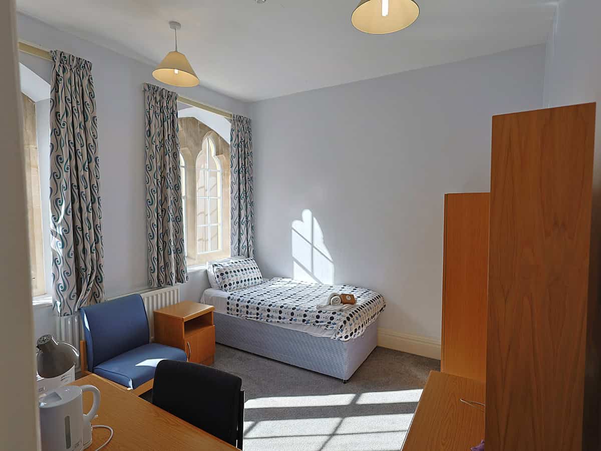 Bright single bedroom with a bed, desk, chairs and wooden wardrobes, lit by sunlight through tall Gothic-style windows with patterned curtains.